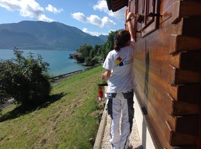 Mitarbeiterin beim Streichen der Holzfassade mit See im Hintergrund